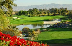 Golf course on a lake with red flowers in the foreground and trees in the background.